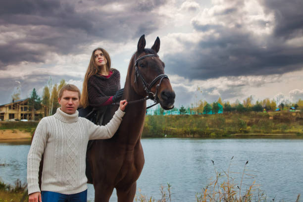 Young couple riders with horse walking in fall forest by lake, looking at camera. People equestrian man and woman on horseback in autumn park. Leisure activity and resting concept. Copy ad text space jóvenes jinetes de pareja con caballos caminando en el bosque de otoño junto al lago, mirando a la cámara. gente ecuestre hombre y mujer a caballo en el parque de otoño. concepto de actividad lúdica y descanso. copiar el espacio de texto del anuncio