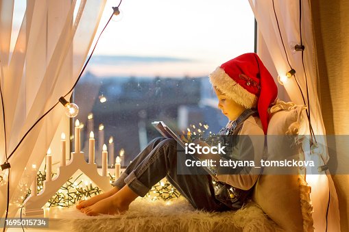 Toddler child, cute blond boy, sitting on the window, reading book, christmas lights around