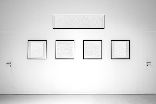 In the white-walled exhibition room, frames with empty interiors are displayed. The white interior of the frames allows the insertion of images or text. Frames aligned between two doors.