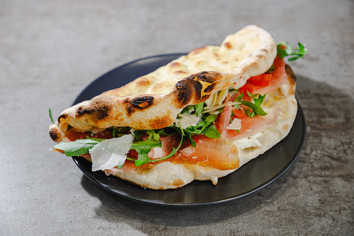 Folded fluffy flatbread sandwich with delicious ingredients prosciutto, tomato slices, grana cheese and arugula, served on a small plate