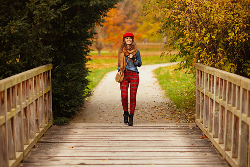 Hello autumn. Full length portrait of smiling trendy woman in jeans shirt and red hat with autumn leaf, scarf and bag walking in the city park.