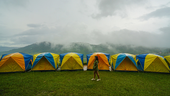 Women walk past camping Tent with the spectacular view of Khao Kho, Phetchabun Province in Thailand. Tent in camping with flog and mountain view. Tent on campsite by the hill in rainy day.