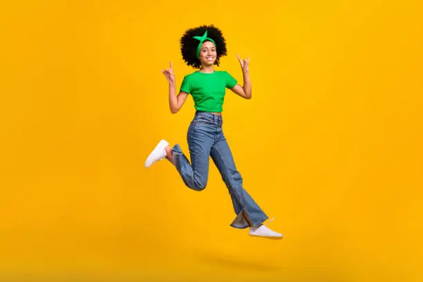Full body portrait of beautiful excited teen schoolgirl jump hands fingers show heavy metal symbol isolated on yellow color background.