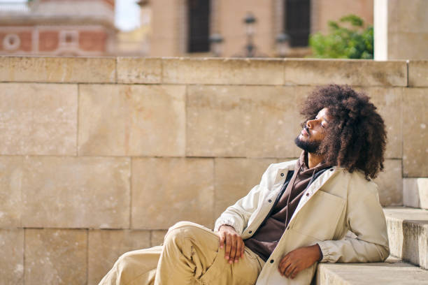 afro man sitting in a square enjoying the sun on his face concept of wellbeing and inner peace stock photo
