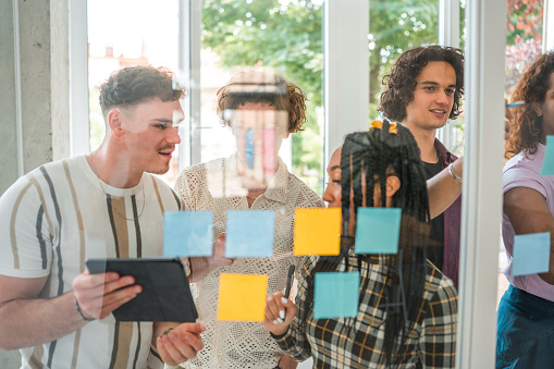 Collective effort as a diverse Gen Z team converges to brainstorm and propel their digital marketing projects forward. Sticky notes cover a glass wall, serving as the canvas for their shared vision, emphasizing the importance of teamwork in the digital age.