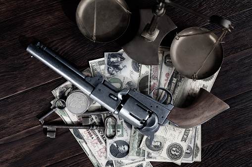 Old west Colt revolver and US banknotes and silver certificates with silver dollar coins and bronze traditional balance scale on wooden table background