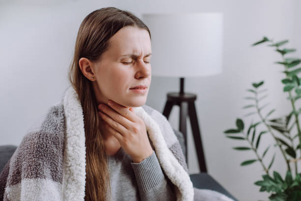 Symptoms from flu season in winter concept. Close-up of unhealthy sad young woman covered blanket sitting on grey couch at home suffering from sore throat, angina, hard to swallow, voice loss Symptoms from flu season in winter concept. Close-up of unhealthy sad young woman covered blanket sitting on grey couch at home suffering from sore throat, angina, hard to swallow, voice loss painfully stock pictures, royalty-free photos & images