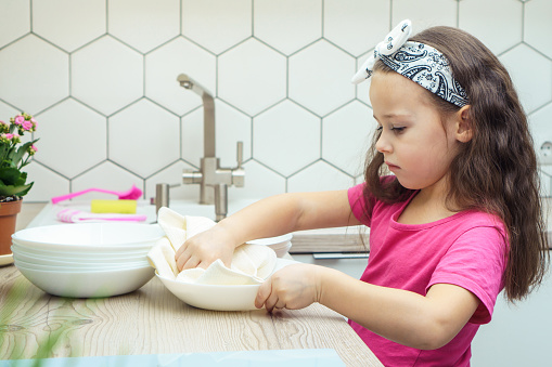 Upset little girl wiping clear bowls with dry towel on kitchen set. Portrait of child washing up dishes in kitchen sink side view. Home cleaning concept. Dry tableware after washing.