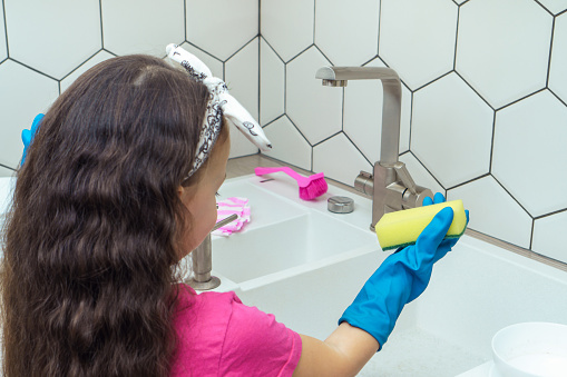 Back view of little girl with long dark hair wearing pink T-shirt, blue rubber gloves, standing near white sink, holding yellow sponge in kitchen, looking down. Dishwashing, housework, housekeeping.