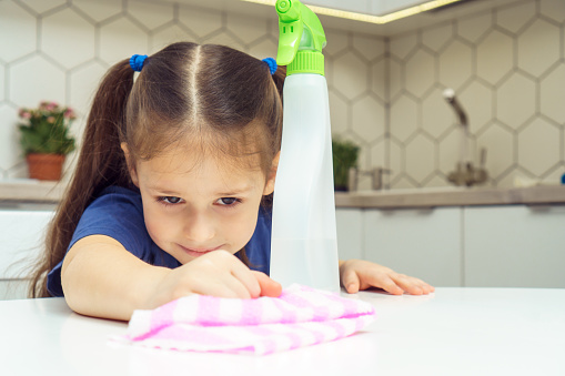 Portrait of smiling little preteen girl with dark hair wearing blue T-shirt cleaning washing white table with rag near spray bottle cleanser in kitchen. Housework, housekeeping, disinfection, hygiene.