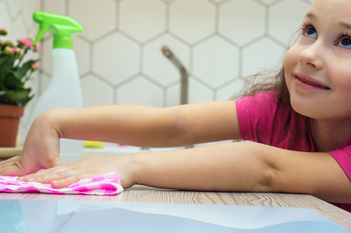 Portrait of joyful little preteen girl with dark hair wearing pink T-shirt, washing cleaning surface of table with rag near spray bottle, looking up in kitchen. Housework, housekeeping, sanitizing.