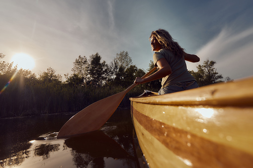 Low angle view of happy woman from the back enjoying while paddling a boat during springtime on a river at sunset. Copy space.