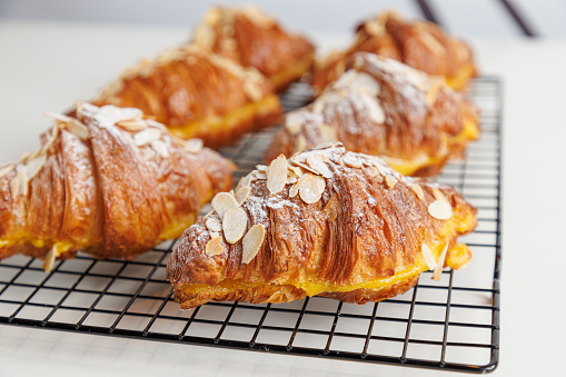Yellowish brown beautifully baked crusty croissant filled with cream, almond flakes topping and powdered sugar icing, served on a grid
