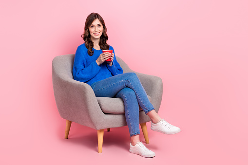 Full size photo of lovely positive girl sit comfy soft chair hold coffee mug empty space isolated on pink color background.