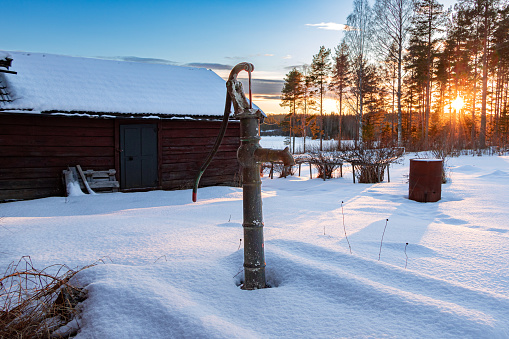 Farm pump in the snow with the sun setting behind the trees in the background