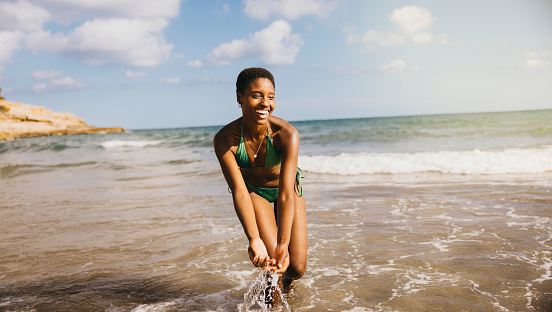 Cheerful smiling black woman in bikini splashing water from the seashore at the beach