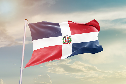 Dominican flag on flagpole on blue sky background. Dominican flag waving in the wind on a background of sky with white clouds. Place for text. 3d illustration.