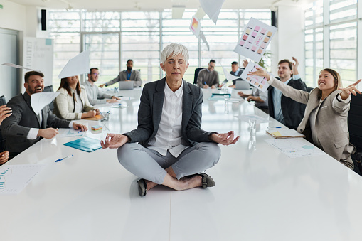 Relaxed senior woman exercising Yoga on the table while her colleagues are throwing reports in the office. Copy space.