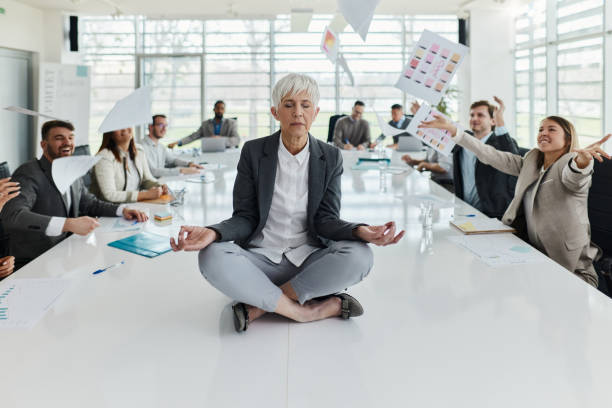 Maintaining mental balance on the job! Relaxed senior woman exercising Yoga on the table while her colleagues are throwing reports in the office. Copy space. chaos stock pictures, royalty-free photos & images
