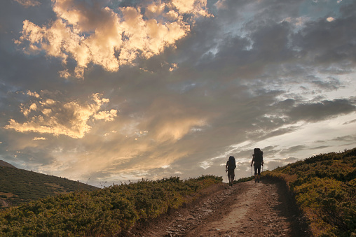 Two hiker silhouettes on the hiking trail high in the mountains. Two men with backpacks and walking poles go towards the horizon in the tcenic sunset mountains.