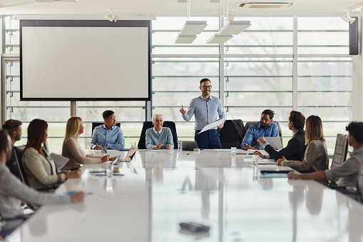 Happy business leader standing in the office and talking to large group of his colleagues during a meeting at conference table. Copy space.