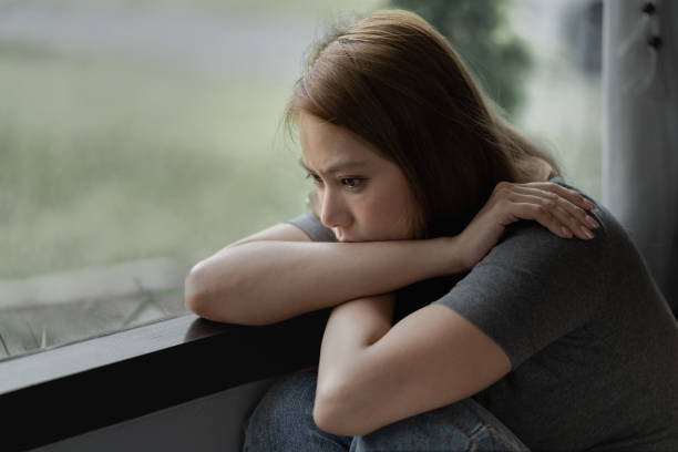 istock Depressed woman sitting by a large window with sad emotion on her face expression 1695317810