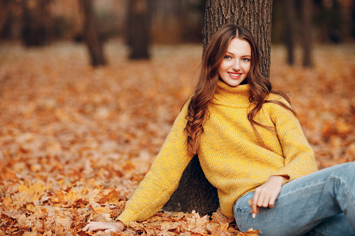 Young smiling woman sitting at tree in autumn park with yellow foliage maple leaves falls.