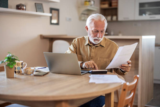 Planning, documents and finance with elderly man on laptop in a kitchen, reading retirement and savings plan in his home. thinking, mortgage and investment paperwork with retired male contemplating.