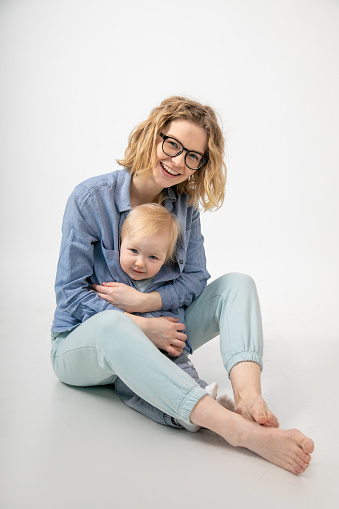 Portrait of beaming family sitting on white background. Young laughing woman mother holding embracing little blond blue-eyed girl baby daughter, covering with shirt, posing. Motherhood, childhood.