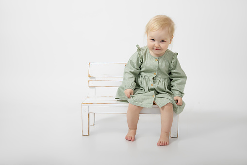 Portrait of smiling little blond barefoot girl with blue eyes wearing green cotton dress, sitting on white wooden little bench on white background, looking at camera. Childhood. Studio, copy space.