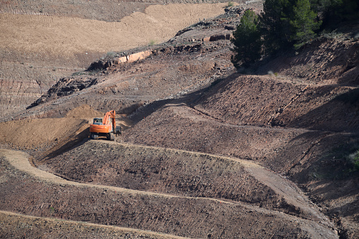 The excavator is orange and in the middle left of the photo. All around it are signs of earth having been dug, and excavations being made. There is a line zig zagging through the photo, indicating a track which is clearly used in the excavations. At the top right, in shadow, are some trees.

The photo was taken at a salt mine in Cardona, in the Spanish region of Catalonia, in the province of Barcelona. The salt mine is part of a mountain mass called Muntanya de Sal. The hill has been worked as a mine since Roman times; pieces cut from it have been carved by artists in Cardona into images, crucifixes and many articles of an ornamental kind. The photo was taken in May 2023