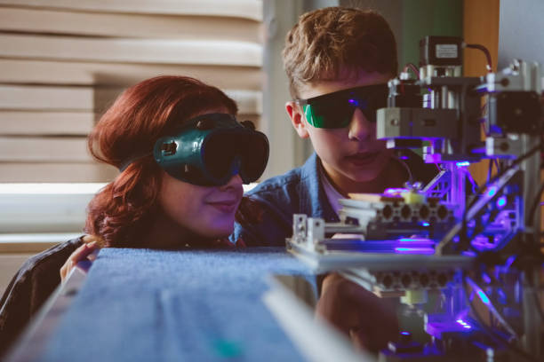 Gen Z teenage girl and boy wearing protective glasses watching laser engraving and cutting machine in contemporary high school laboratory.