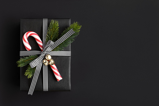 Christmas black gift box with candy cane, bells and spruce twigs on dark background. Copyspace.