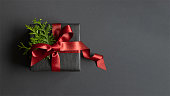 Black gift box with thuja and red ribbon on dark background. Flatly, copyspace.