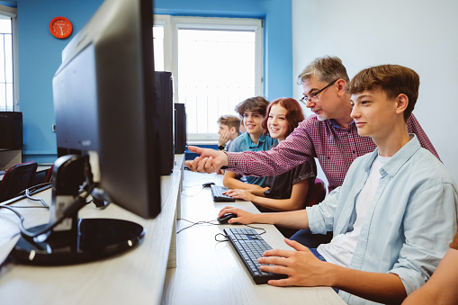 Gen Z teenage high school students coding durning computer lessons, talking with male teacher, sitting in computer lab.
