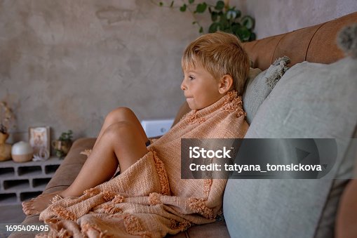 Blond child, boy, sitting on the couch at home,cuddled in blanket, enjoying free time