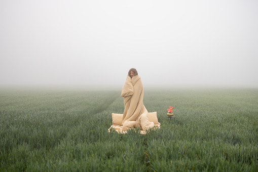Woman wakes up on bed covered with bedsheet on green field during foggy morning. Concept of dreams and freedom