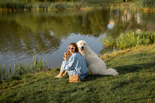 Young woman eats ice cream while sitting with her cute white dog near the lake in park. Friendship with pets and spending leisure time together
