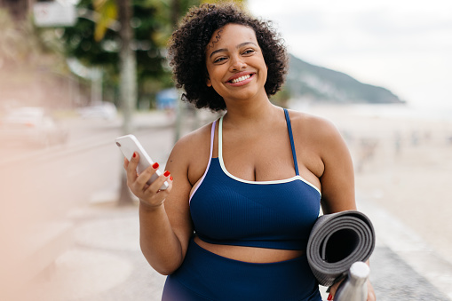 Fit young woman stands on a beach promenade by the ocean, holding a yoga mat and essentials. She's happily smiling while using her smartphone, enjoying her healthy workout routine which includes beach yoga.