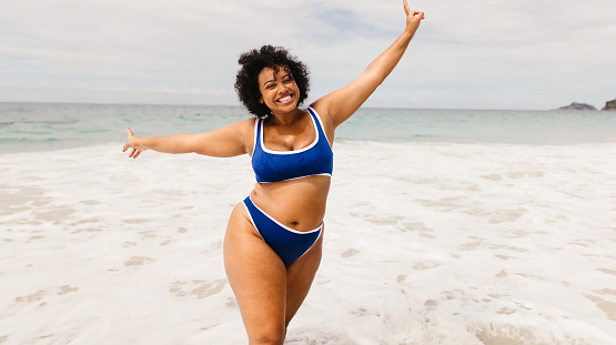 Happy young woman dancing and celebrating at the beach, wearing a bikini and raising her arms in excitement. Carefree, plus size woman having fun on a solo summer vacation.