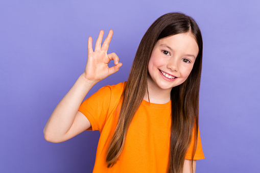 Photo of toothy beaming schoolkid with straight hair dressed orange t-shirt showing okey good work isolated on purple color background.
