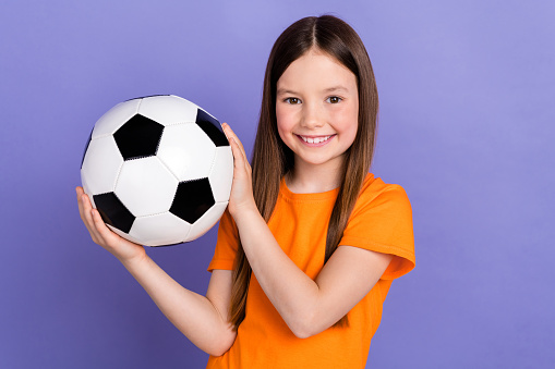 Photo of toothy beaming schoolkid with straight hair dressed orange t-shirt arms hold soccer ball isolated on purple color background.