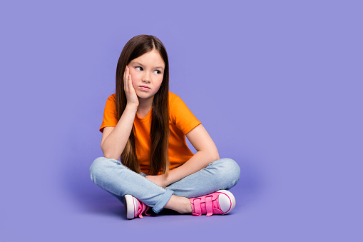 Full size photo of minded sad schoolgirl wear stylish t-shirt denim trousers sit look empty space isolated on violet color background.