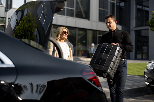 Businesswoman waits while chauffeur packs a suitcase in car trunk, using luxury taxi service during a business trip. Concept of business transfer services, idea of personal driver.