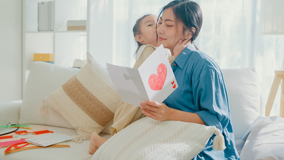 Young Asian family mom and daughter hugging and reading handmade greeting card with heart having fun on sofa in living room during holiday celebration mothers day at home. Family happy moment concept.