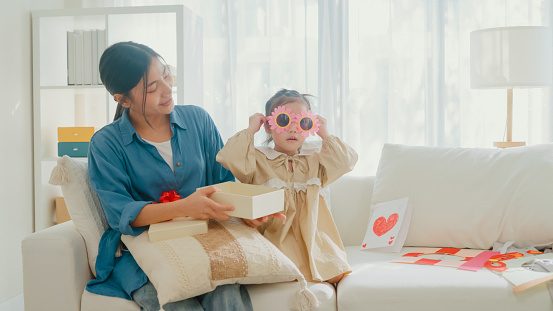 Young Asian family child wearing sunglass make funny face sitting on couch play together in living room during holiday celebration mothers day at home. Family happy moment concept.