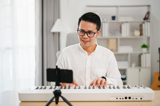 Asian man wearing white headphones play piano and sing a song and learning online with mobile phone and compose writing song or record sound with a microphone and use computer recording music program