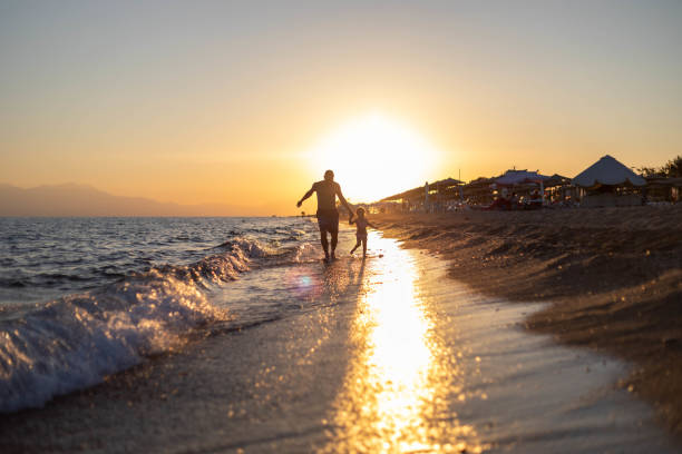 A man runs by the sea with his daughter. A beautiful sunset is behind them, and they enjoy the waves of the sea lapping them as they run.