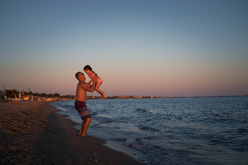 A man and his little girl are playing on the seashore. The child is delighted, because the father holds her high and spins her.