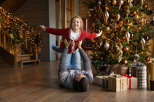 Happy young father lying on warm heated floor, lifting in air adorable little child daughter, having fun near decorated Christmas tree in stylish living room, New Year holidays family celebration.